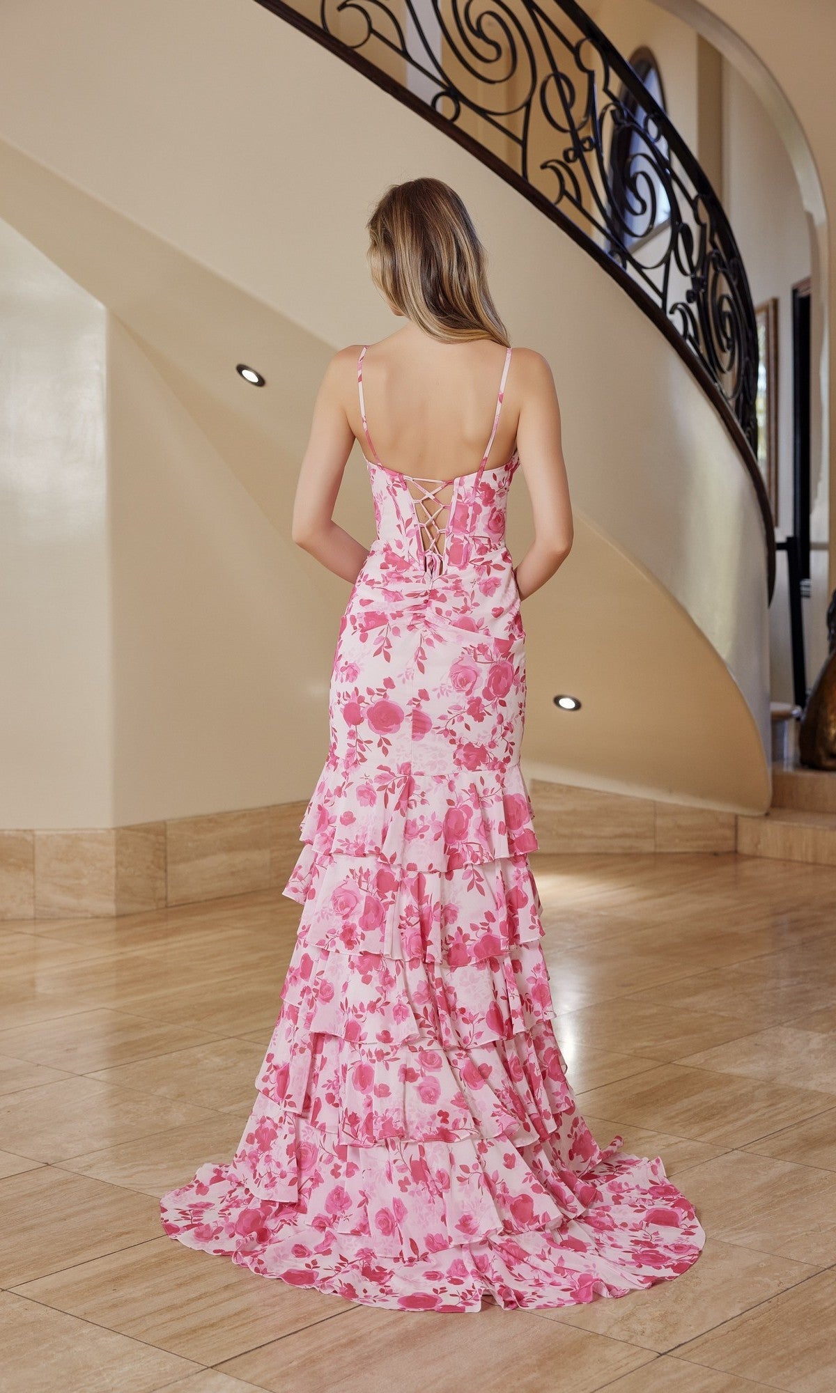 A woman stands indoors in the Nox Anabel T1863, a pink and white floral chiffon mermaid prom dress with a backless design, her elegant gown flowing as she faces a curved staircase.